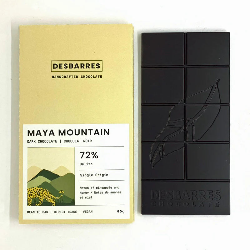 This image displays a bar of Desbarres Maya Mountain 72% dark chocolate and its accompanying pale yellow packaging, presented side-by-side against a clean white background.
The primary subject is a bar of dark chocolate, presented as a rectangular slab divided into eight individual squares. The chocolate is a deep, rich black, suggesting a high cocoa content. Embossed on its surface is a stylized depiction of a cacao pod or leaf, a common motif for chocolate products. Below the embossed design, the brand n
