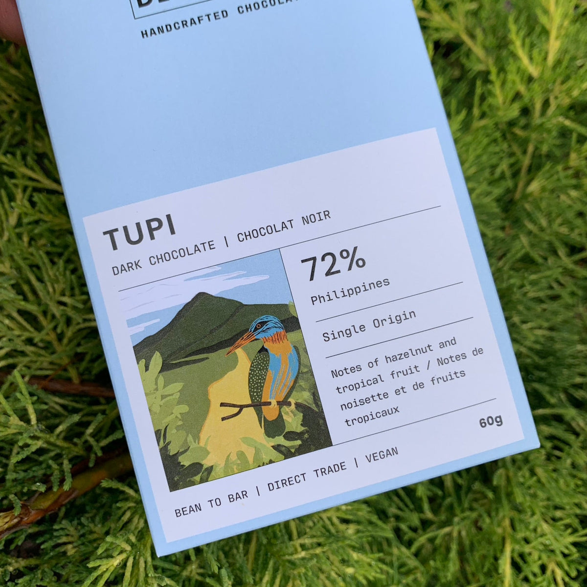 A light blue chocolate bar wrapper displaying an illustration of a kingfisher bird on a branch with a mountainous landscape in the background. Text on the wrapper indicates "TUPI", "DARK CHOCOLATE", "72%", "Philippines", "Single Origin", "Notes of hazelnut and tropical fruit", "60g", and "BEAN TO BAR | DIRECT TRADE | VEGAN".


